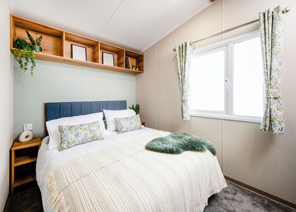 Een slaapkamer met een bed, een houten plank met planten, een raam met gordijnen en een nachtkastje met een lamp.