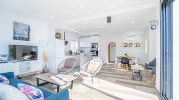 Una sala de estar moderna con sofá, mesa de centro y sillas, una zona de cocina con armarios y electrodomésticos, y un comedor con mesa y sillas.