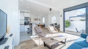 A modern living room with a sofa, two armchairs, a coffee table, and a TV. There is a kitchen area with a dining table and chairs, and a large window offering a view of the outside. - Villa in Empuriabrava with Private Pool (Empuriabrava)