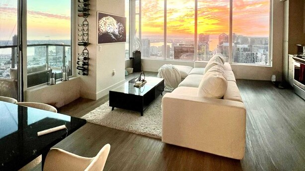 A modern living room with a large sofa, a coffee table, and a dining area with chairs. There are large windows offering a view of the city skyline at sunset. - Penthouse High-rise Ocean City Views (San Diego)