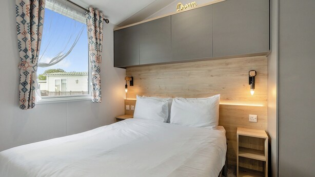 Quarto de hotel com cama, mesas de cabeceira, janela com cortinas estampadas, cabeceira com iluminação embutida e vista para um edifício.