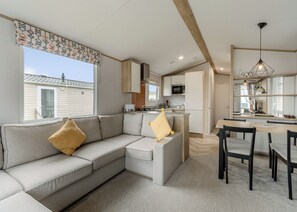 Ein modernes Wohnzimmer mit grauem Sofa, einem Esstisch mit Stühlen, einer Küchenzeile mit Schränken und Geräten sowie einem großen Fenster mit gemusterten Vorhängen.