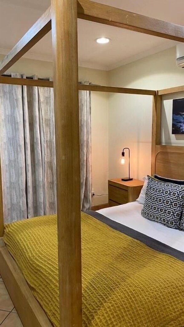 Una habitación con una cama de dos columnas de madera, una mesita de noche con lámpara y un cojín estampado.