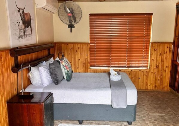 Una cama con cabecera de madera, una mesita de noche, un ventilador de pared y una ventana con persianas.