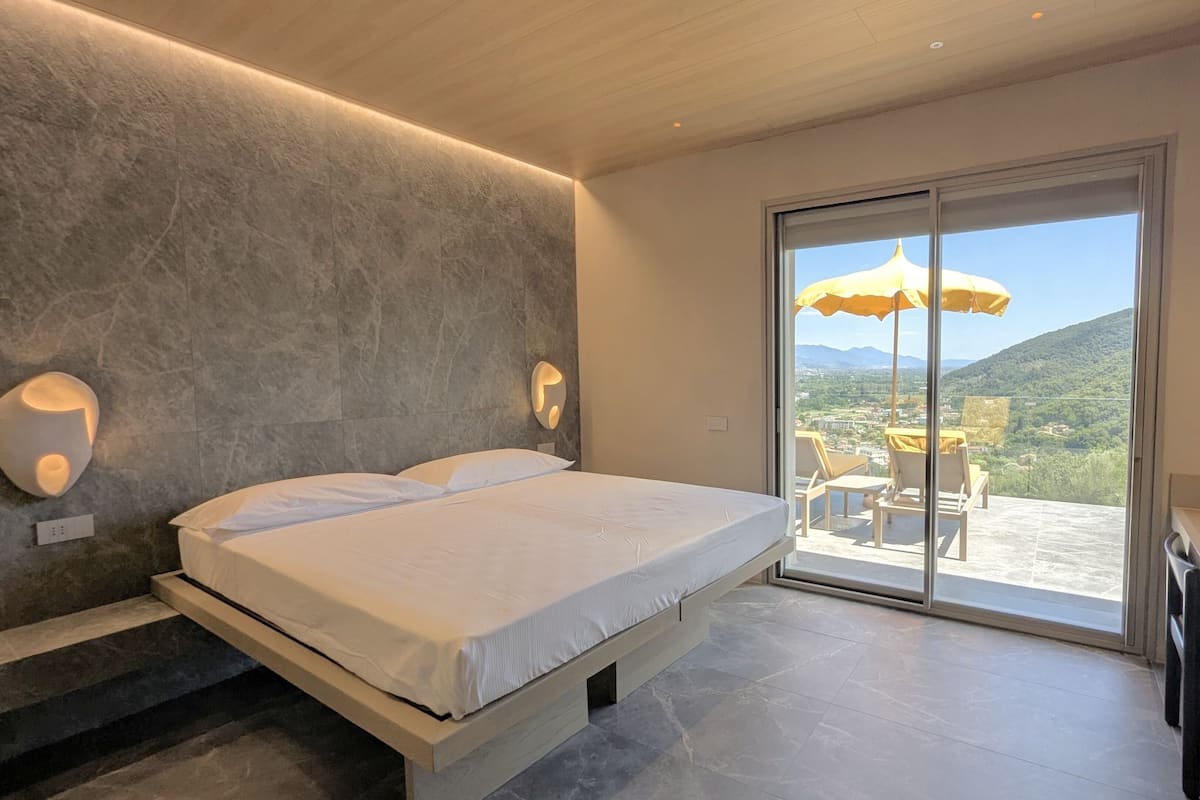 Un dormitorio moderno con una cama grande, un balcón con una sombrilla amarilla y vistas a la ciudad.