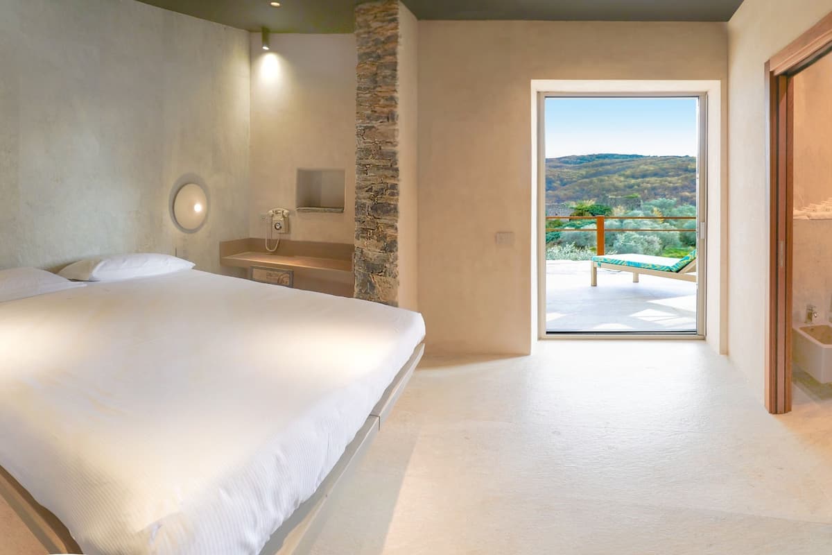 Un dormitorio con una cama grande, un pilar de piedra, una mesita de noche, un teléfono y vistas a un balcón con una silla.