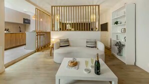 Una sala moderna con un sofá blanco, una mesa de centro con una botella y vasos, y una zona de cocina con encimera y armarios de madera.