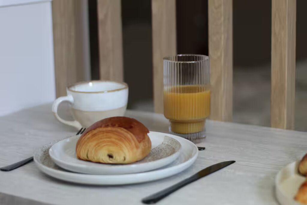 En morgenmadsopsætning med en croissant på en tallerken, en kop kaffe og et glas appelsinjuice.