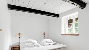 Ein kleines Zimmer mit einem Einzelbett, weißen Wänden, Holzboden, einem Fenster mit Blick auf Grünflächen und einem schwarzen Balken an der Decke.