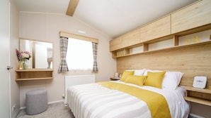 A bedroom with a bed, a bedside table, a mirror, a window with curtains, a radiator, and a wooden headboard. - 2 bedroom accommodation in Talacre, Holywell (Talacre, Holywell)