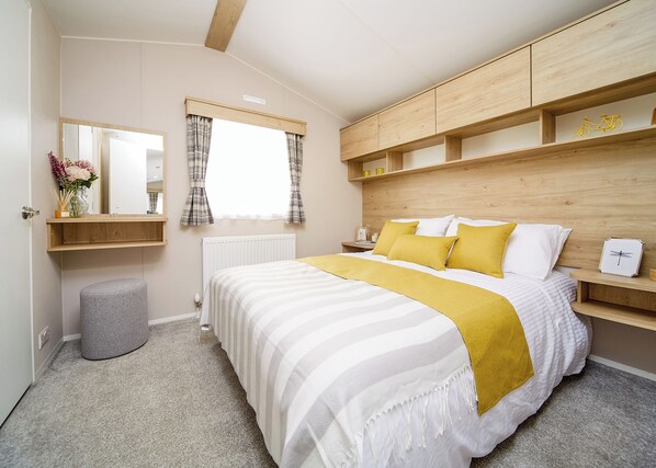 A bedroom with a bed, a bedside table, a mirror, a window with curtains, a radiator, and a wooden headboard. - 2 bedroom accommodation in Talacre, Holywell (Talacre, Holywell)