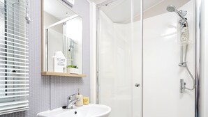 A bathroom with a glass shower enclosure, a white sink with a mirror above it, and a window with blinds. - 2 bedroom accommodation in Talacre, Holywell (Talacre, Holywell)
