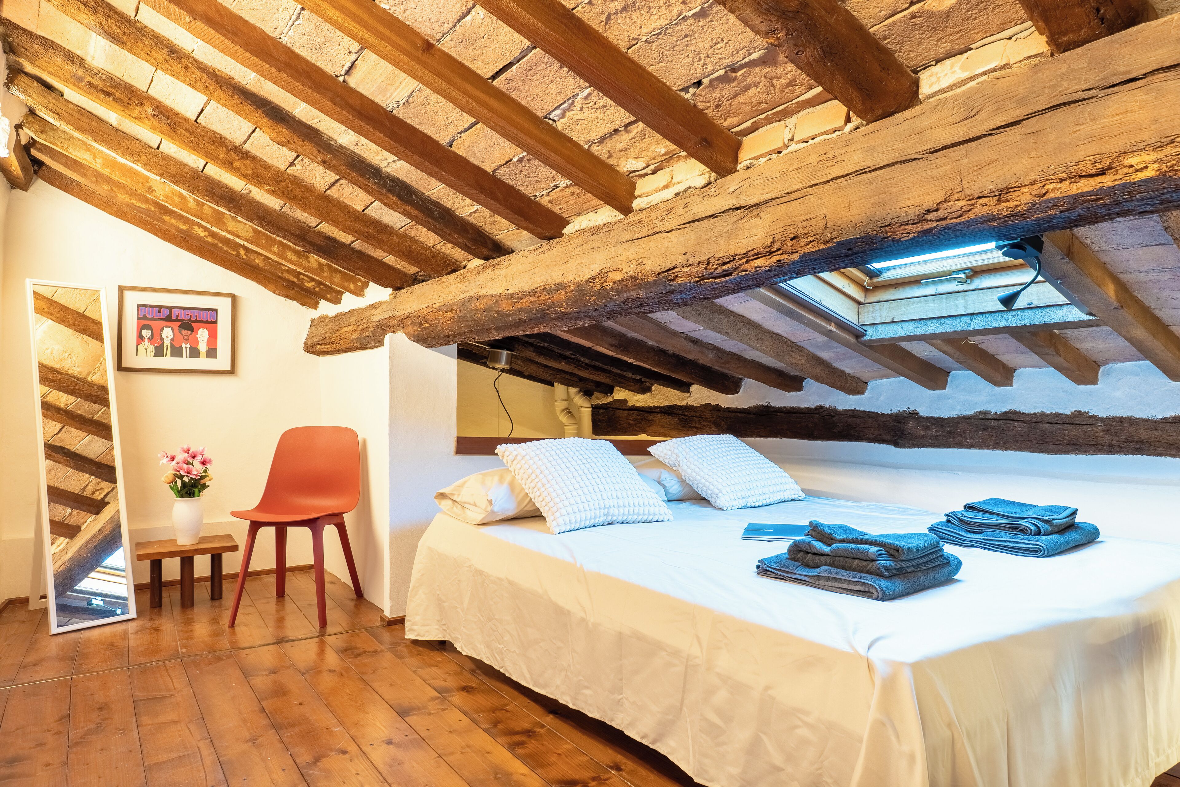 A bedroom with a wooden ceiling, a bed with white bedding, a red chair, and a small table with a vase of flowers.