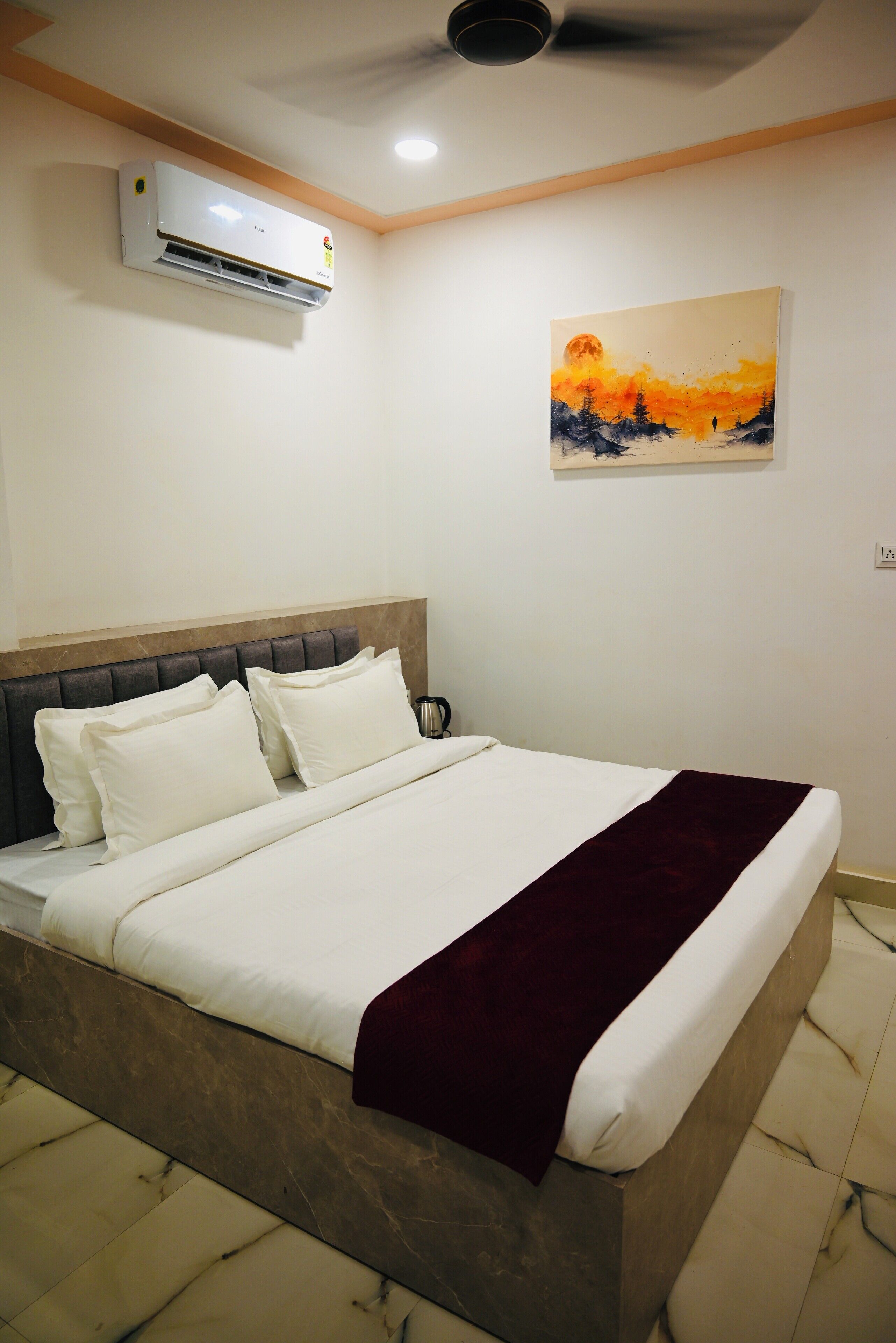 Ein Schlafzimmer mit einem Bett, weißer Bettwäsche, einem dunklen Kopfteil, einer roten Tagesdecke, einer Klimaanlage und einem Bild an der Wand.