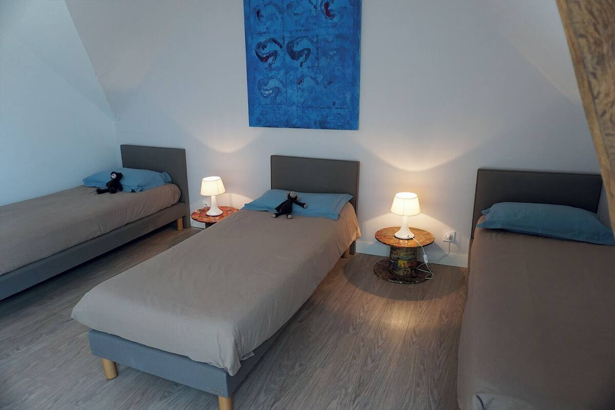 A room with three beds, each with a headboard and a bedside table with a lamp. A blue abstract painting is on the wall above the beds.