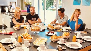 Une table dressée pour le petit-déjeuner, avec un groupe de personnes assises autour, et divers aliments présentés, notamment des viennoiseries, des fruits et des boissons.