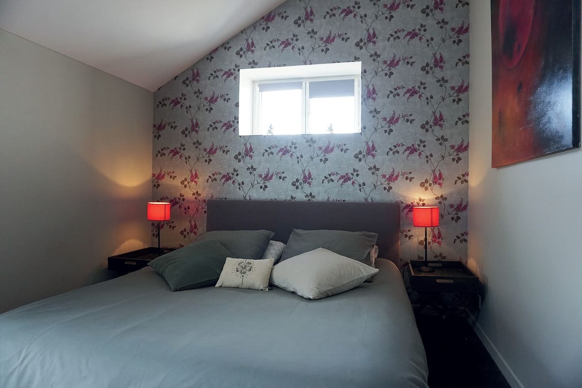 A bedroom with a bed, two bedside lamps, a window, and floral wallpaper.