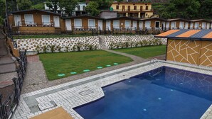 A swimming pool with a tiled deck, a grassy area, and a row of small buildings with doors and windows. - Fillow Casa Hills (Narendra Nagar)
