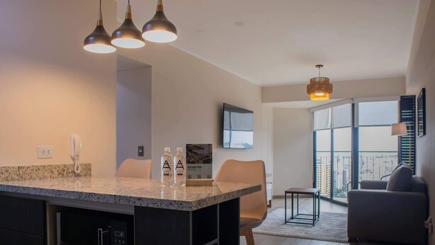 A modern kitchen with granite countertops, a built-in oven, and a coffee machine. There is a dining area with a table and chairs, and a living room with a sofa and a TV mounted on the wall. - w* | Awesome 2BR Apartment in Miraflores (Lima)