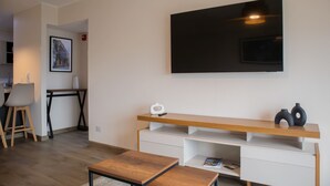A modern living room with a flat-screen TV mounted on the wall, a wooden coffee table, a white entertainment unit, and a dining area with a table and chairs. - w* | Awesome 2BR Apartment in Miraflores (Lima)