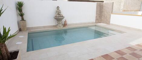 Una piscina con una estatua y una pequeña fuente, rodeada por un muro blanco y plantas en macetas.