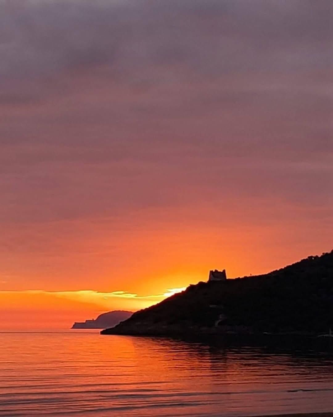 A sunset over a coastal landscape with a silhouette of a hill and a structure on it.