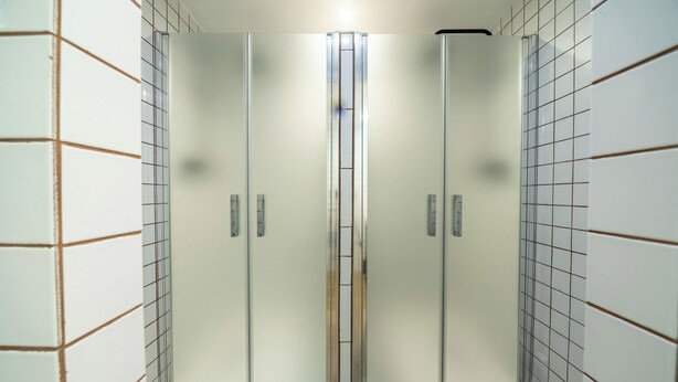 Une armoire à portes coulissantes avec un miroir, dotée d’une cloison centrale et flanquée de deux portes, dans une pièce dont les murs sont recouverts de carreaux et le sol est en bois.