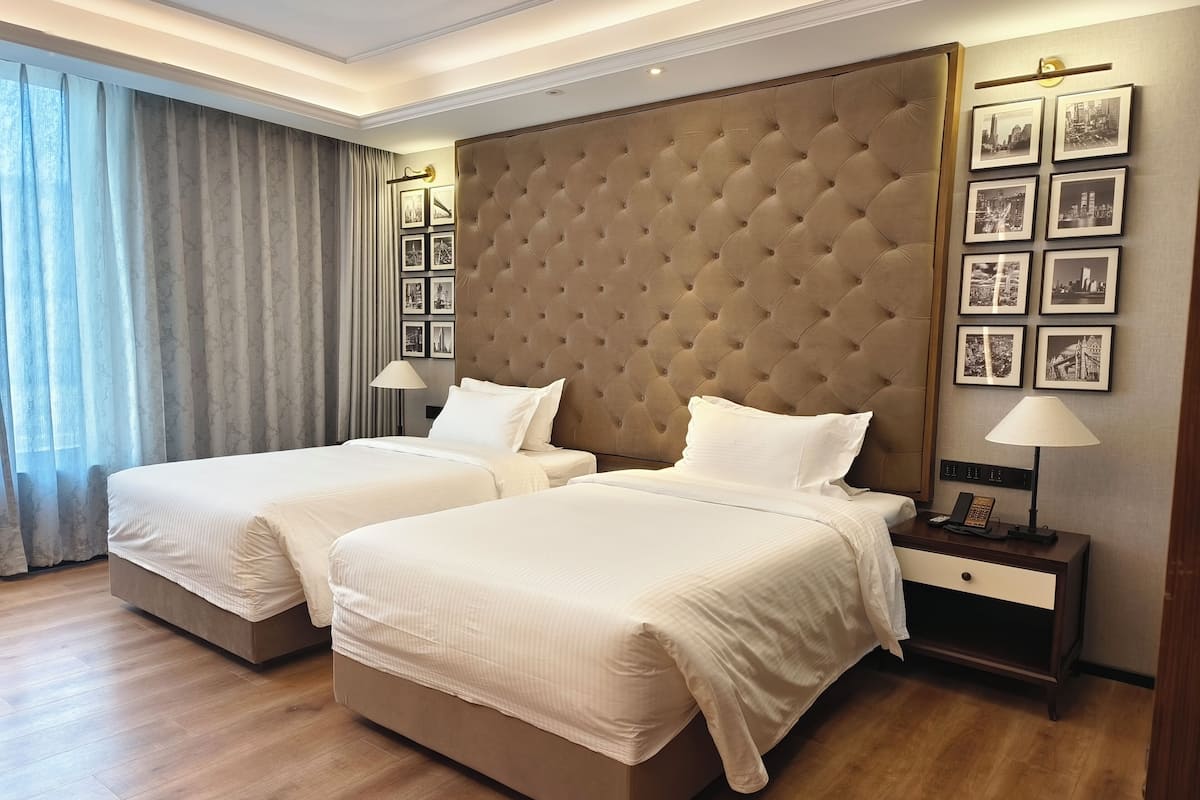 Habitación de hotel con dos camas, un cabecero grande con capitonado, una mesita de noche con lámpara y cuadros en la pared.
