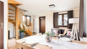 Una sala de estar moderna con una escalera de madera, un comedor con una mesa dispuesta para cuatro personas y un sofá con cojines estampados.