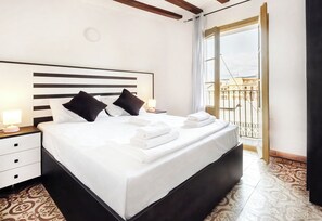 Une chambre d’hôtel avec un grand lit, deux tables de chevet avec des lampes, une vue sur un balcon doté d’une balustrade et un sol recouvert de carreaux décoratifs.