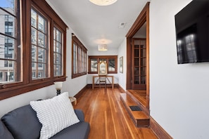A hallway with wooden flooring, a dark-colored sofa with a white cushion, and a large mirror with a wooden frame.