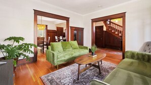 Uma sala de estar espaçosa com um sofá verde, uma mesa de centro de madeira e uma planta grande. Há uma escada que leva a outro andar e uma área de refeições visível ao fundo.