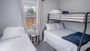 A compact bedroom with a bunk bed, a single bed, a small table, and a window with curtains.