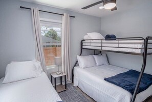 A compact bedroom with a bunk bed, a single bed, a small table, and a window with curtains.