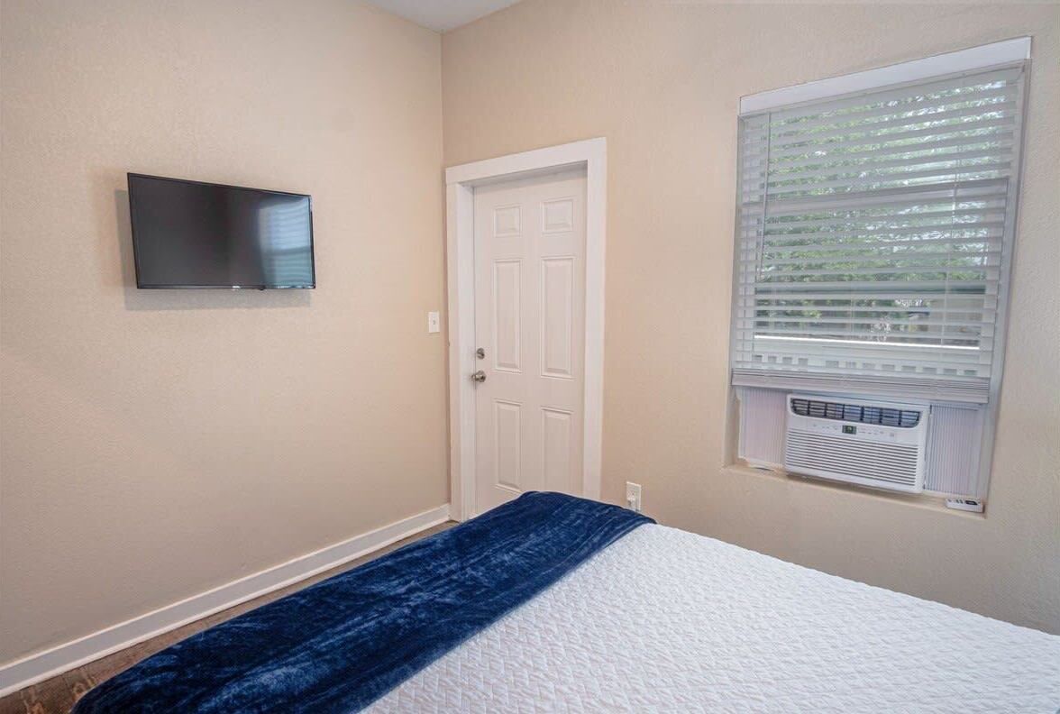 A bedroom with a bed, a TV mounted on the wall, a door, and a window with blinds.