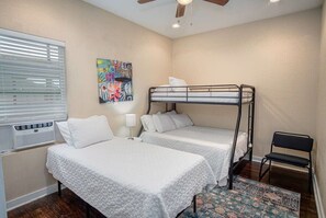 A room with two bunk beds, a window with blinds, an air conditioning unit, a lamp, a chair, and a colorful rug.