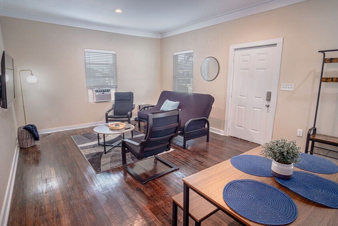 A living room with a dark wood floor, a round coffee table, two black armchairs, a small table with a lamp, and a white door.