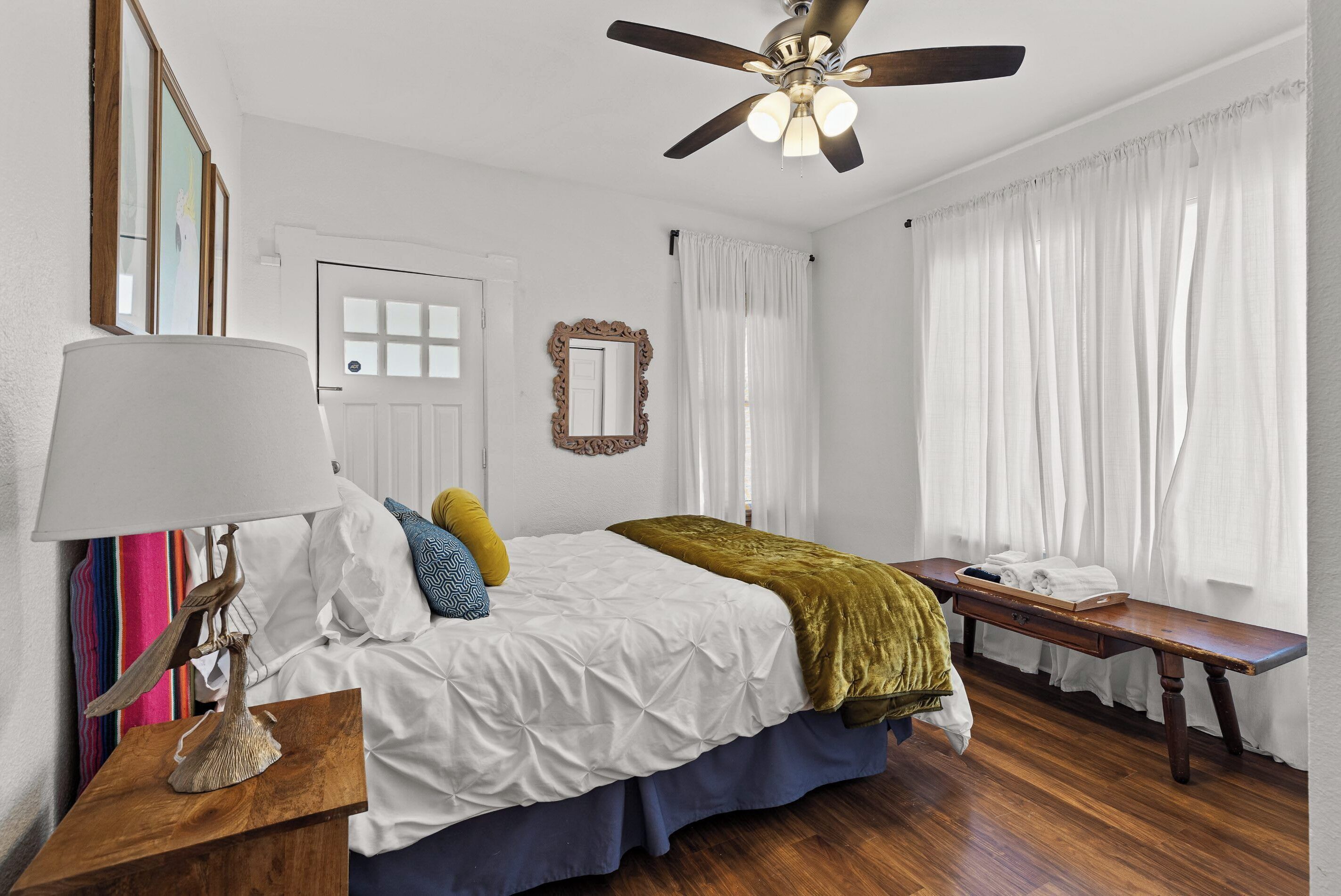A bedroom with a bed, bedside table, lamp, ceiling fan, window with curtains, and a bench by the window.