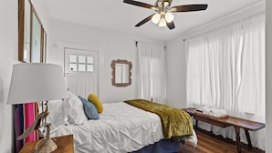 A bedroom with a bed, bedside table, lamp, ceiling fan, window with curtains, and a bench by the window. - Gorgeous Artistic Fully Fenced Home Near Downtown (San Antonio)