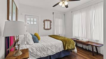 A bedroom with a bed, bedside table, lamp, ceiling fan, window with curtains, and a bench by the window.