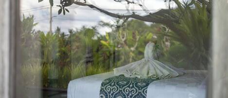 A bed with a patterned blanket, a wooden nightstand, and a view of greenery through a window.