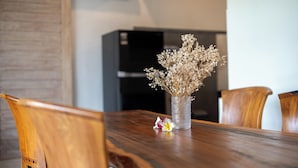 A wooden dining table with a vase of dried flowers and a single flower on it. There are wooden chairs around the table. A black appliance is in the background. - Kesuma Inn Private Villa (Ubud)