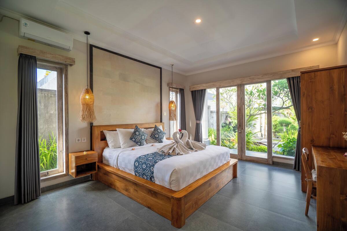 A spacious bedroom with a large bed, wooden headboard, and a view of greenery through large windows.