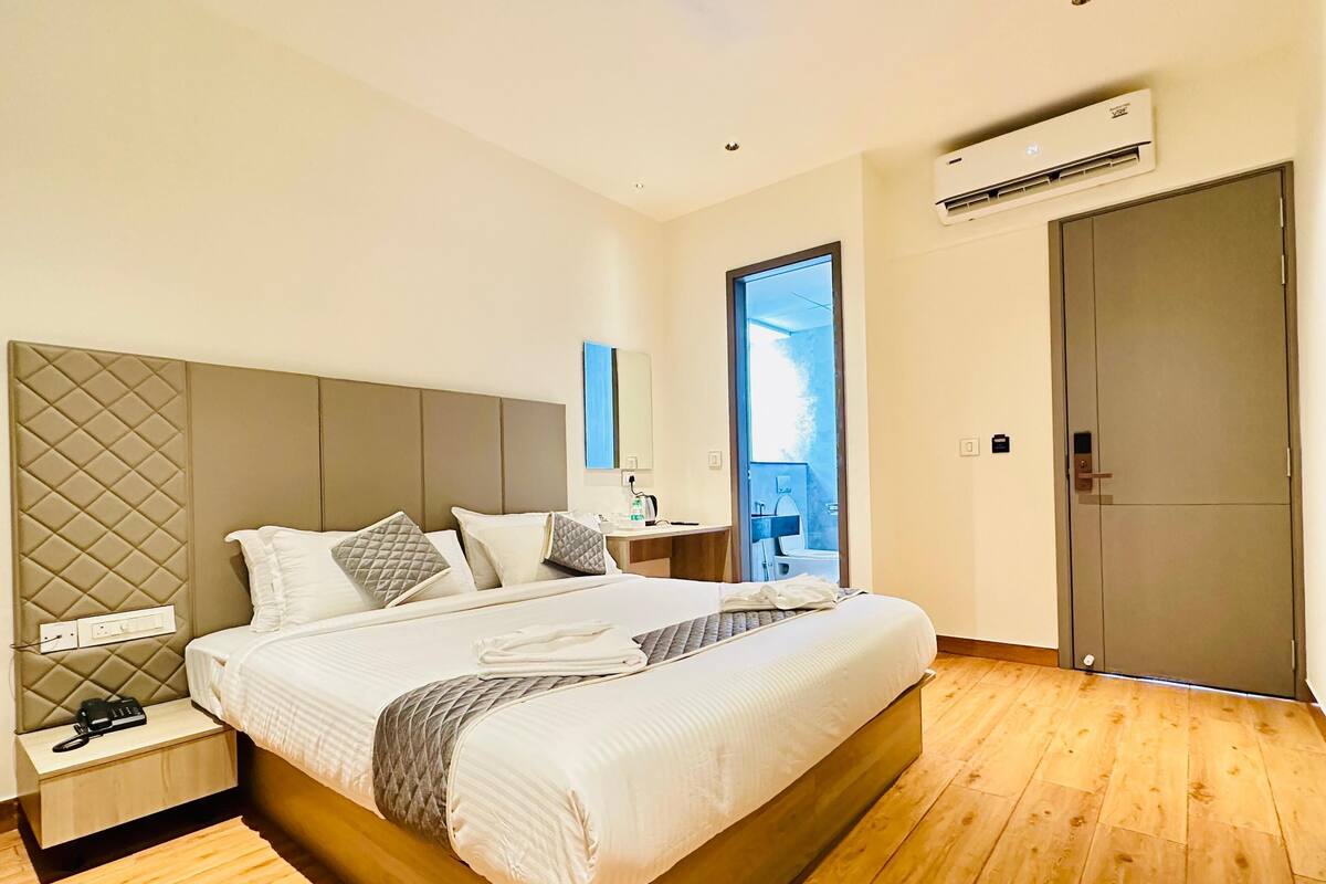 A modern hotel room with a large bed, a bedside table with a phone, a nightstand, an air conditioning unit, a bathroom visible through a glass door, and a wardrobe.