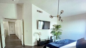 Quarto com cama, televisão, planta e corredor que dá para outro cômodo.