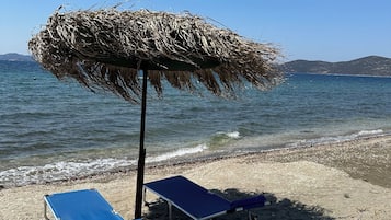 A beach with two blue lounge chairs under a thatched umbrella, a sandy shore, and calm sea in the background.
