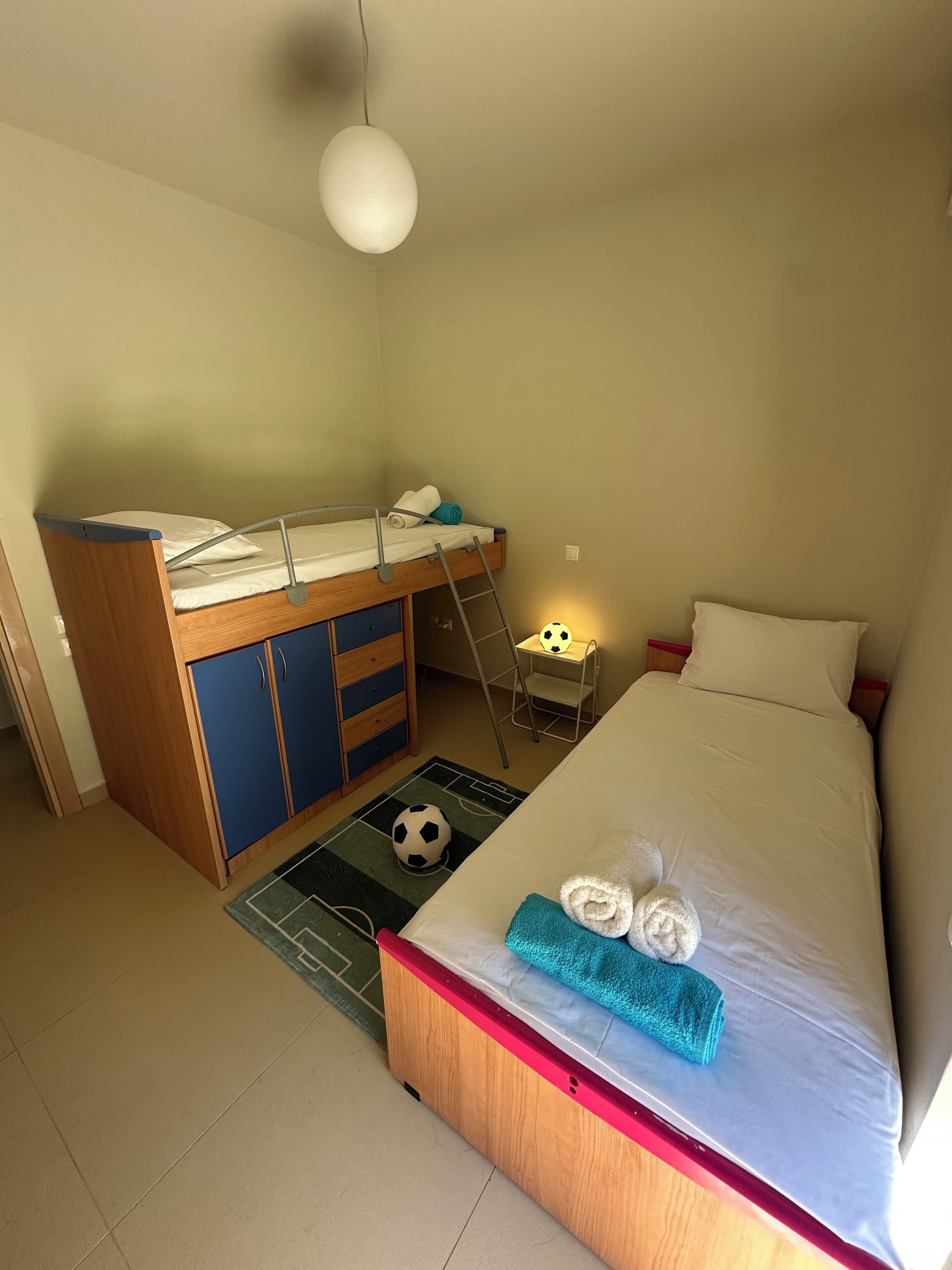 Habitació compacta amb un llit superposat, un escriptori petit, un faldell temàtic de futbol i tovalloles ordenades sobre el llit.