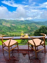 Une table avec deux chaises et un plateau sur lequel se trouvent un verre et une petite statue, le tout donnant sur une vallée verdoyante et luxuriante, parsemée de collines.