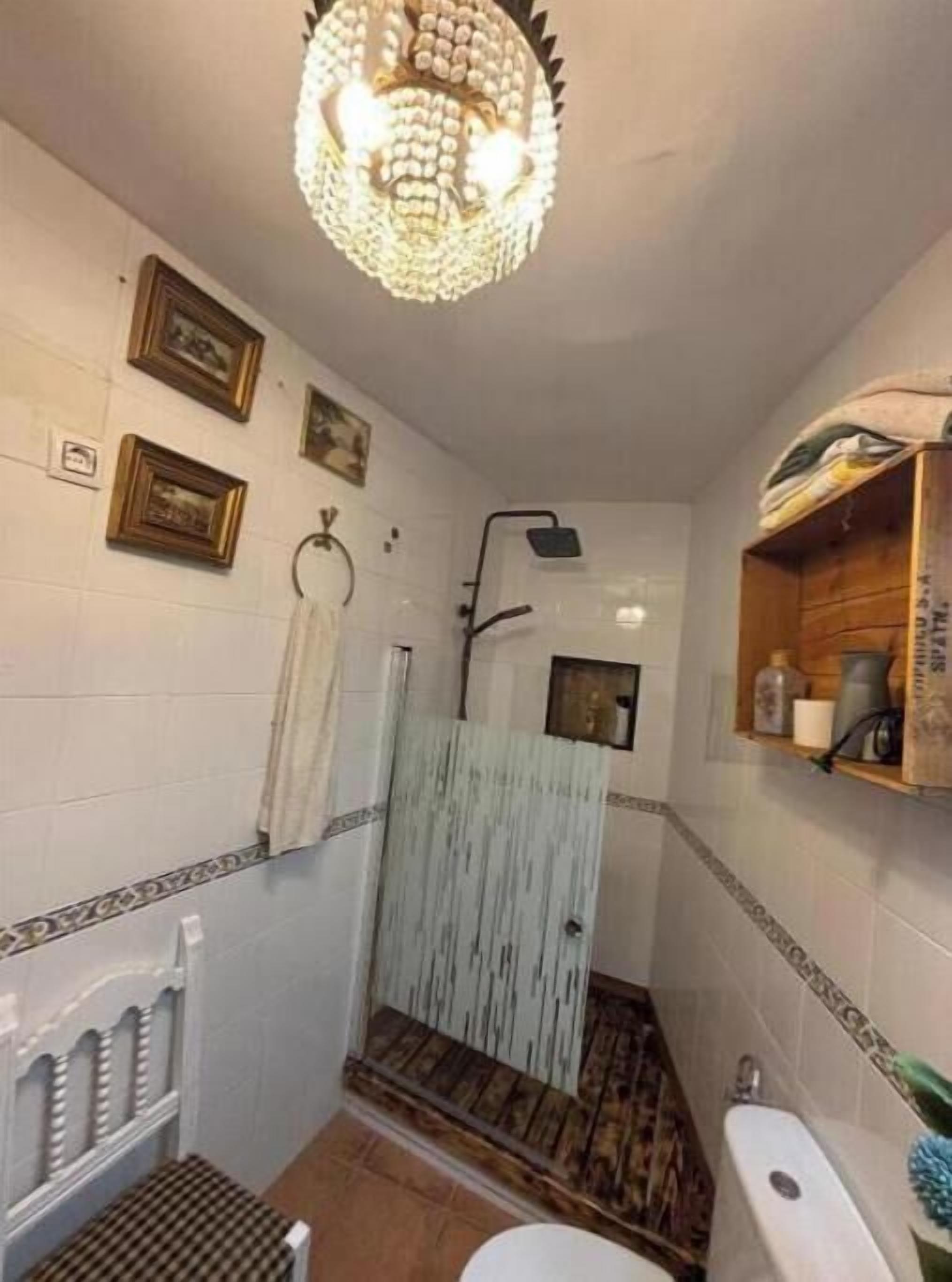 A bathroom with a shower, toilet, and sink. There are framed pictures on the wall and a chandelier above the shower.