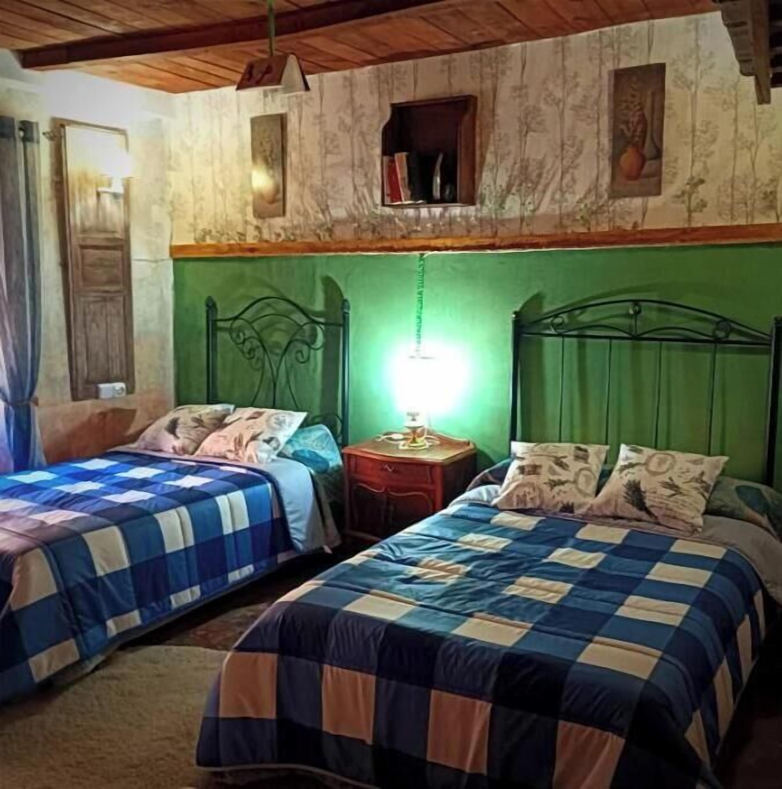 Ein Zimmer mit zwei Betten, jeweils mit einer blau-weiß karierten Bettdecke, einem grünen Kopfteil und einem kleinen Nachttisch mit Lampe.